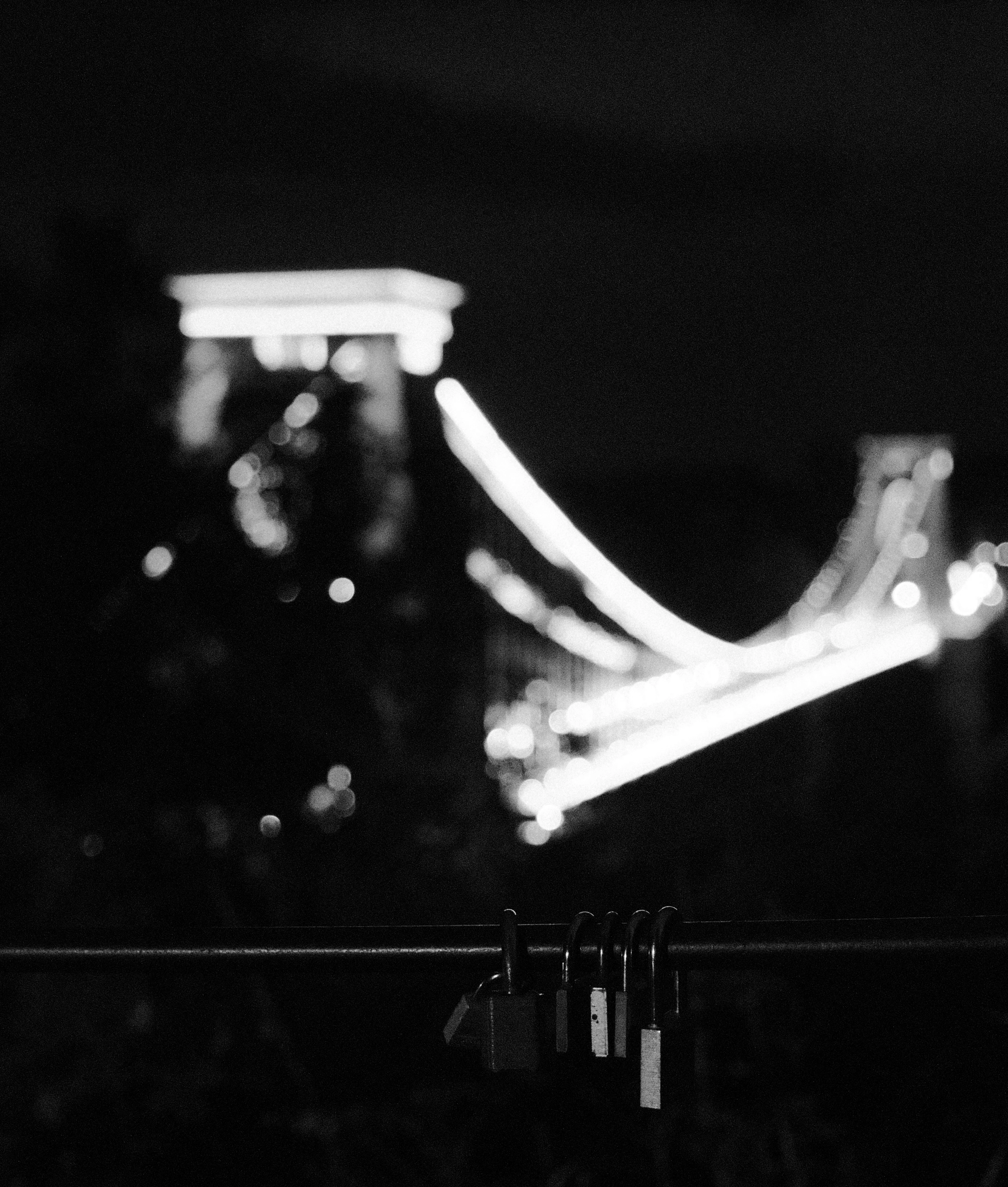 Black and white. Padlocks clipped to a railing, with the lit curve of a suspension bridge blurred behind them.