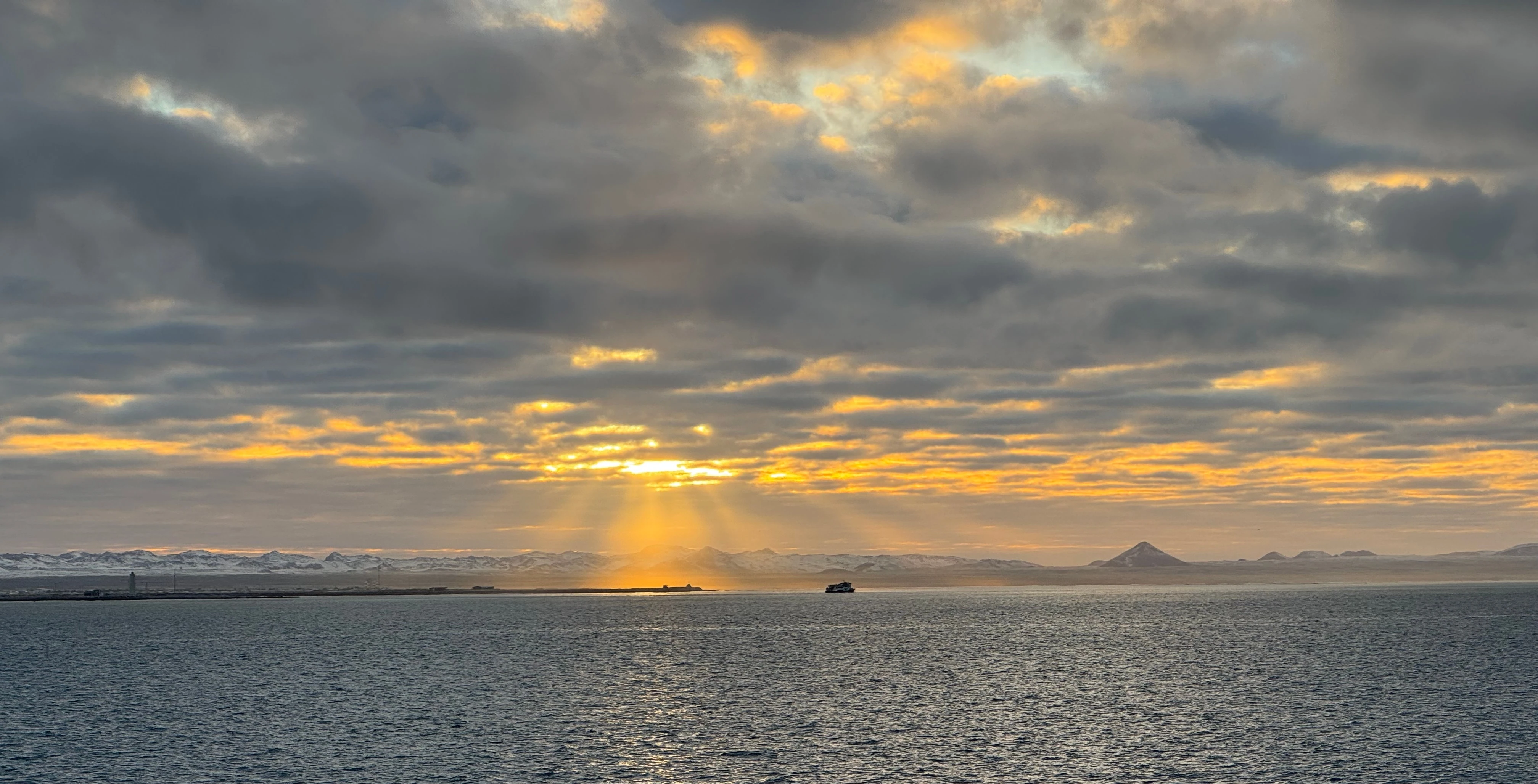 Sunlight breaking through cloud over cold water, with a small ship and low snowy hills on the horizon.