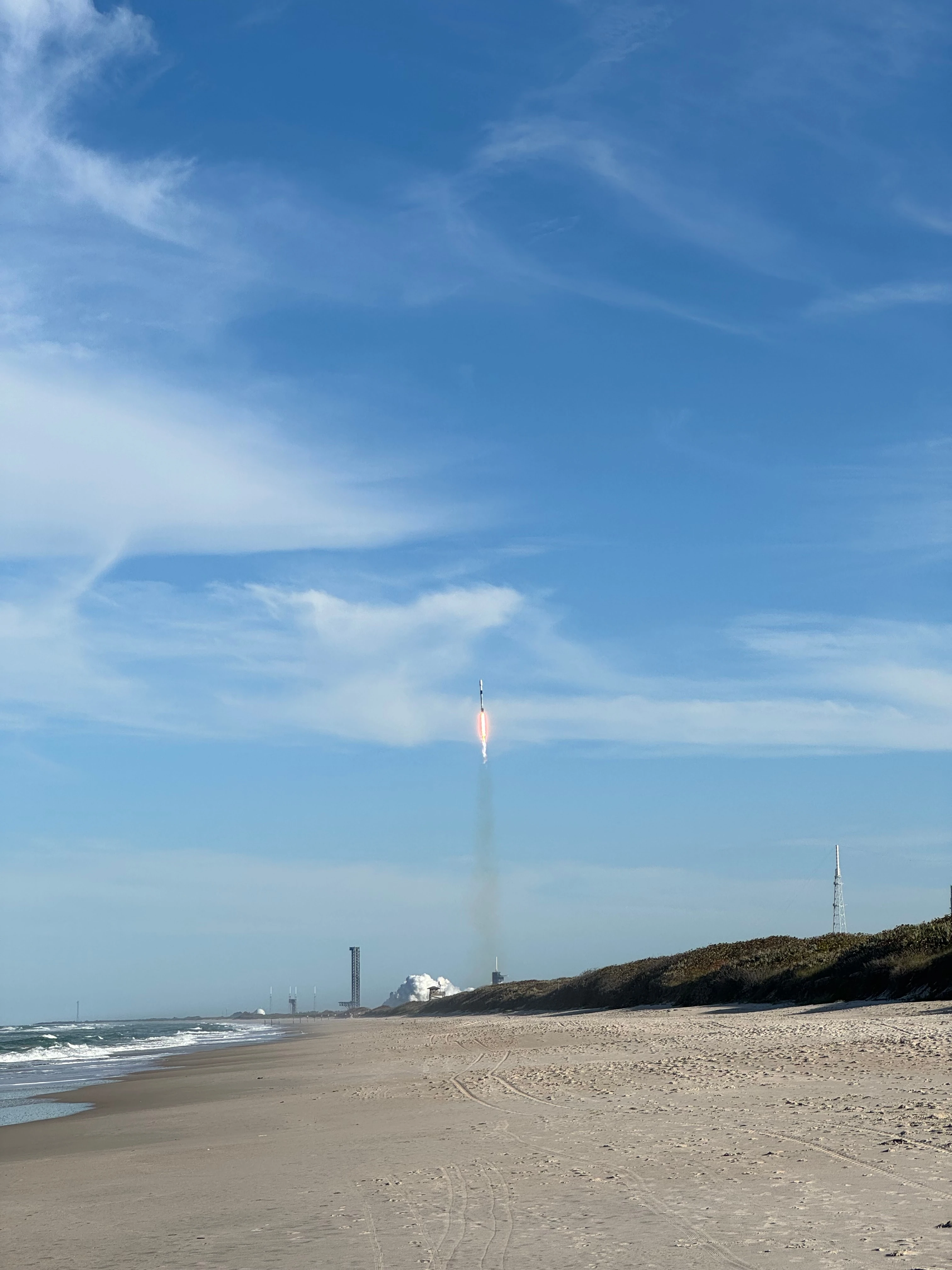 An empty beach with a rocket climbing on a bright plume, leaving a smoke trail up a blue sky.