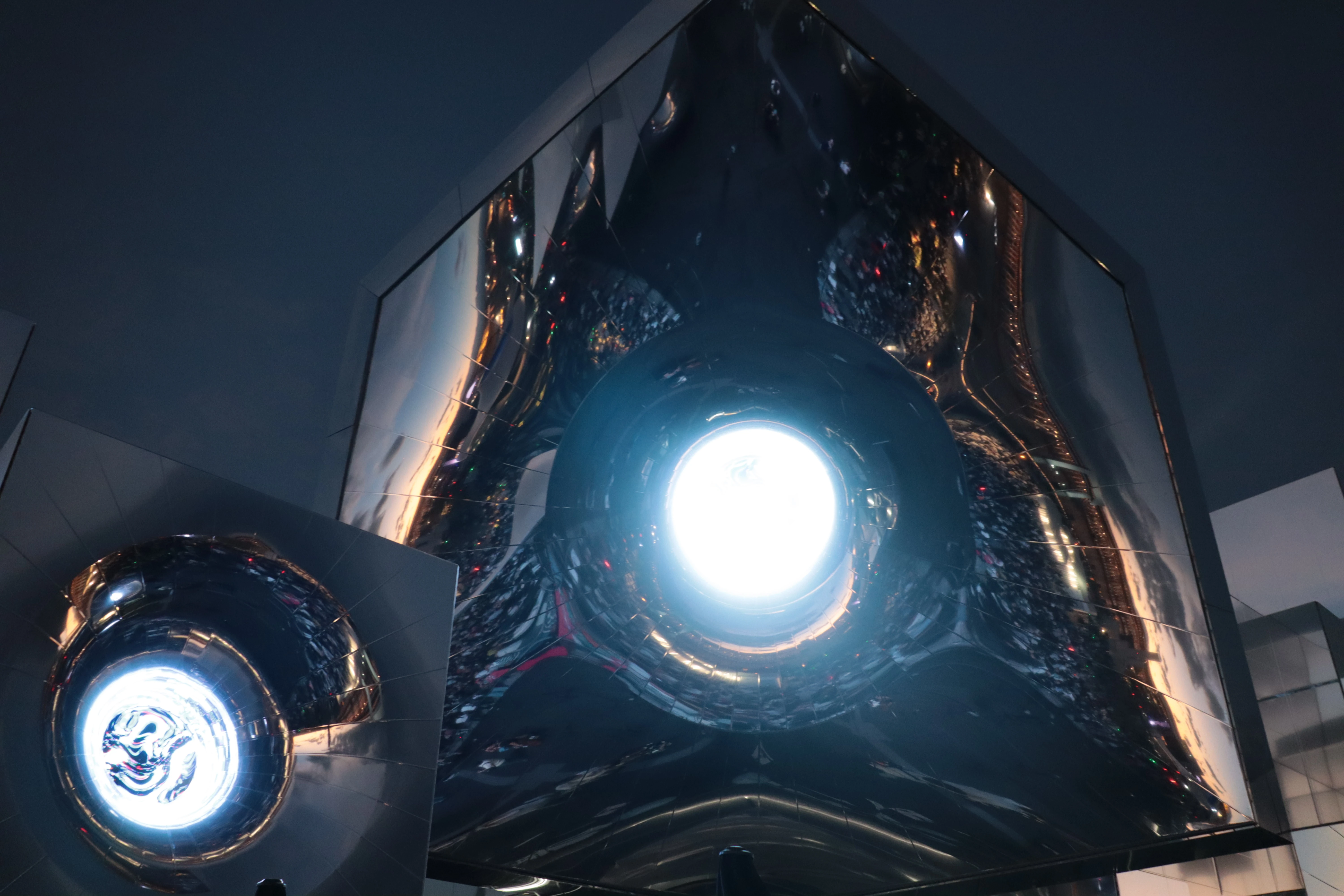 A polished chrome sculpture with a bright circular light at its centre, reflecting the room around it.
