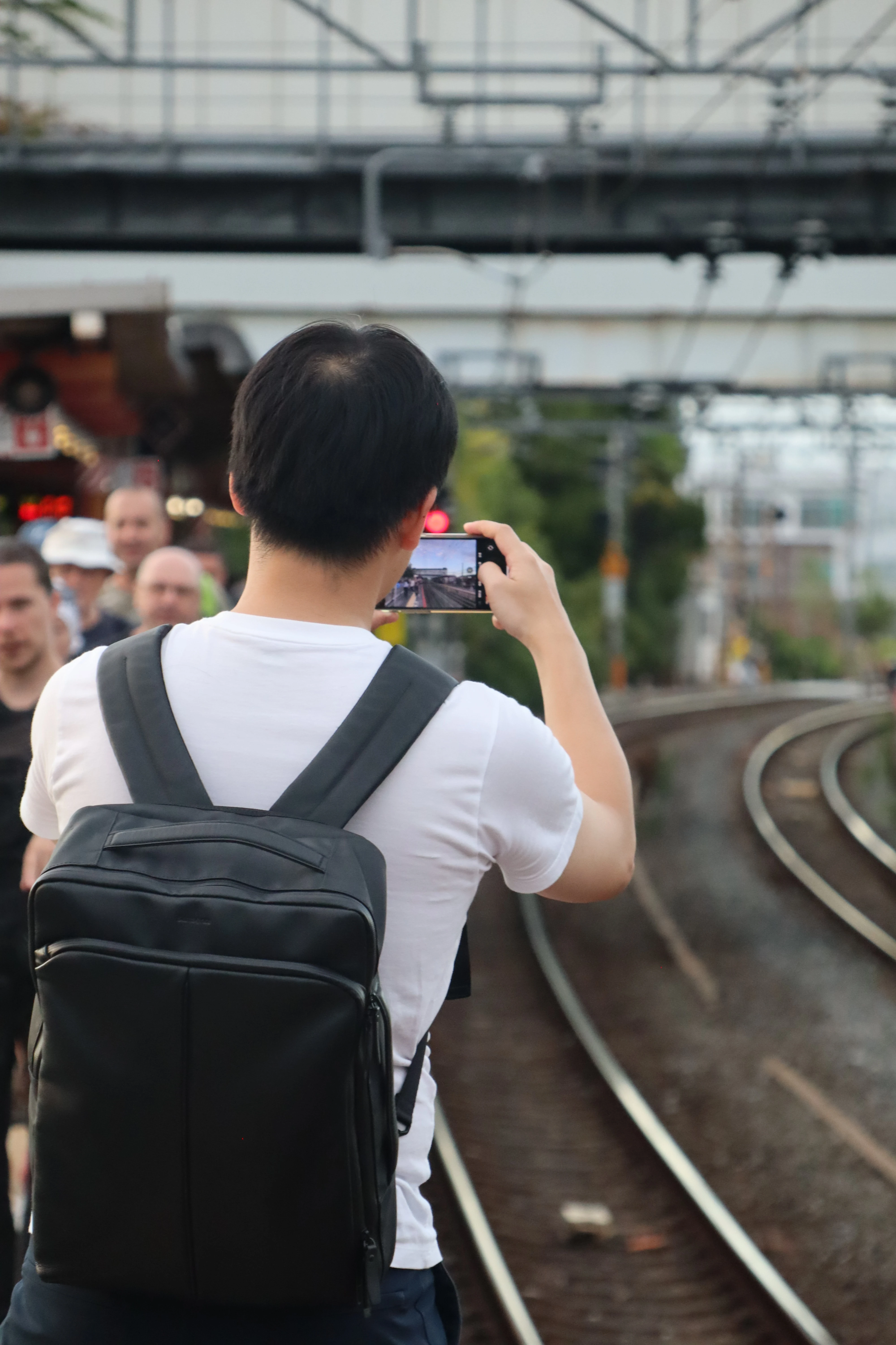 A man seen from behind on a station platform, holding up a phone that shows the railway curving away in front of him.