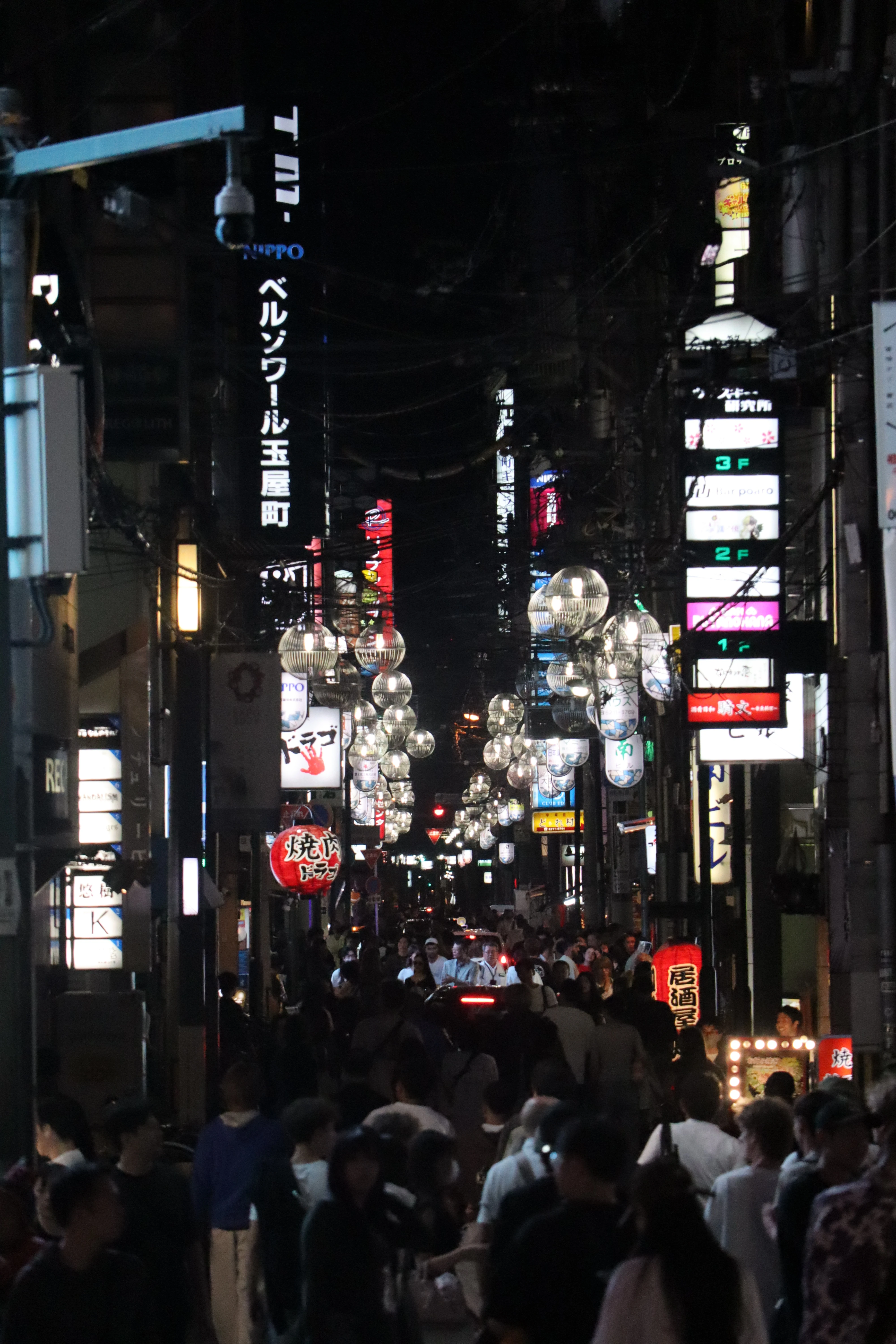 A narrow street at night packed with people, hung with paper lanterns and stacked with illuminated signs.