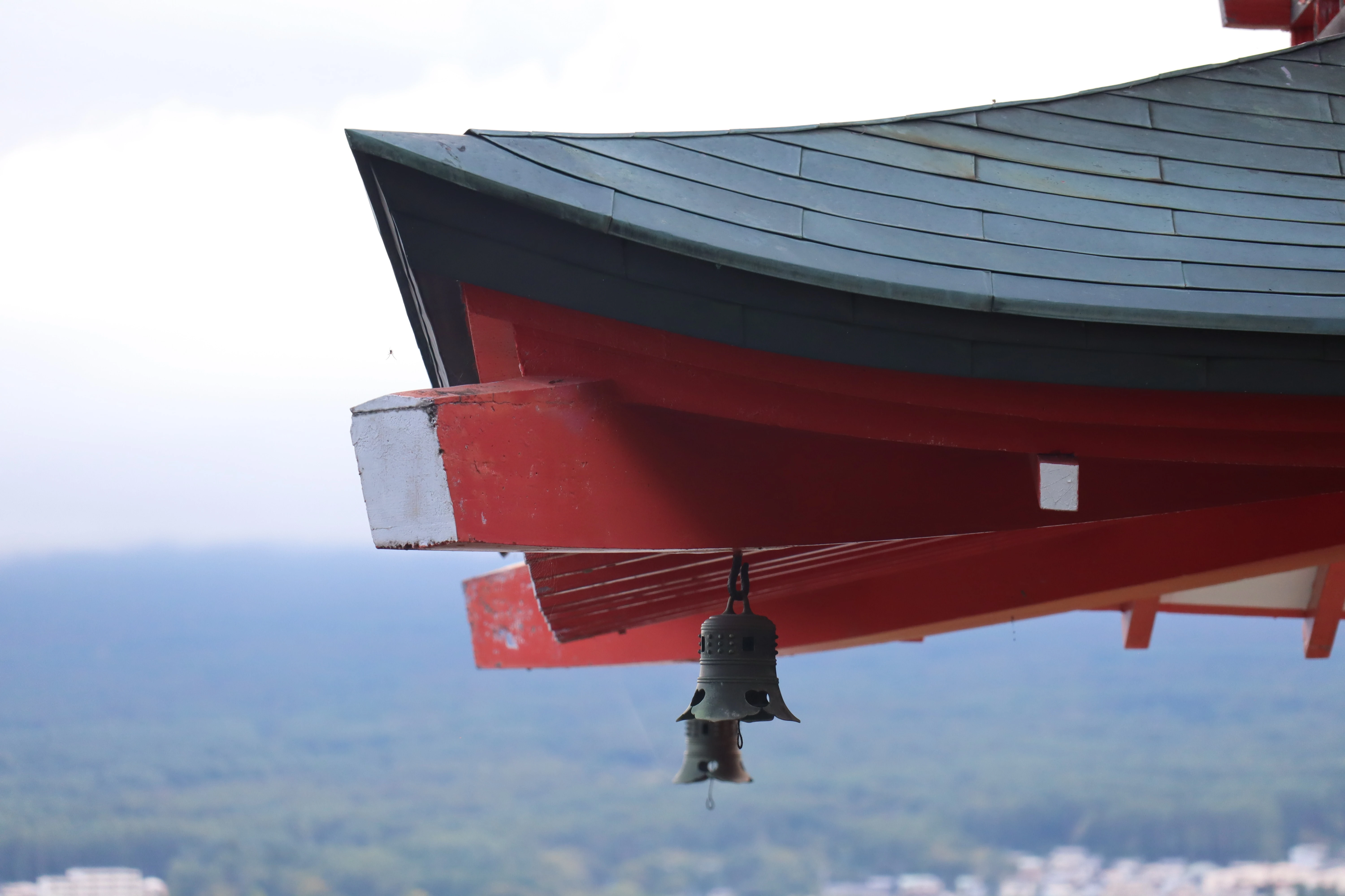 The upswept eave of a red and green temple roof, with a small bronze bell hanging from it against a pale sky.