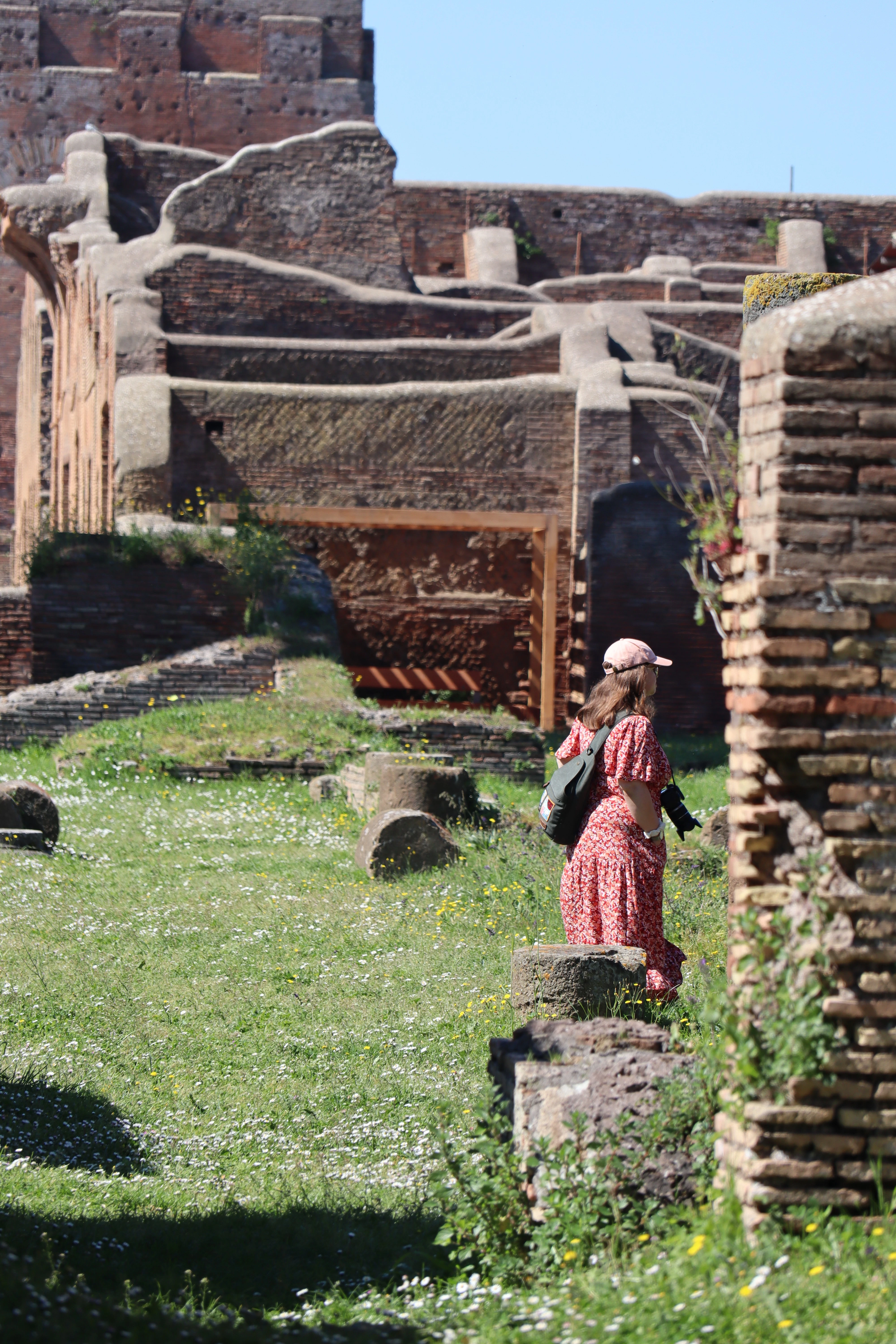 A woman in a long red dress standing among grassed-over Roman brick walls in bright sun.