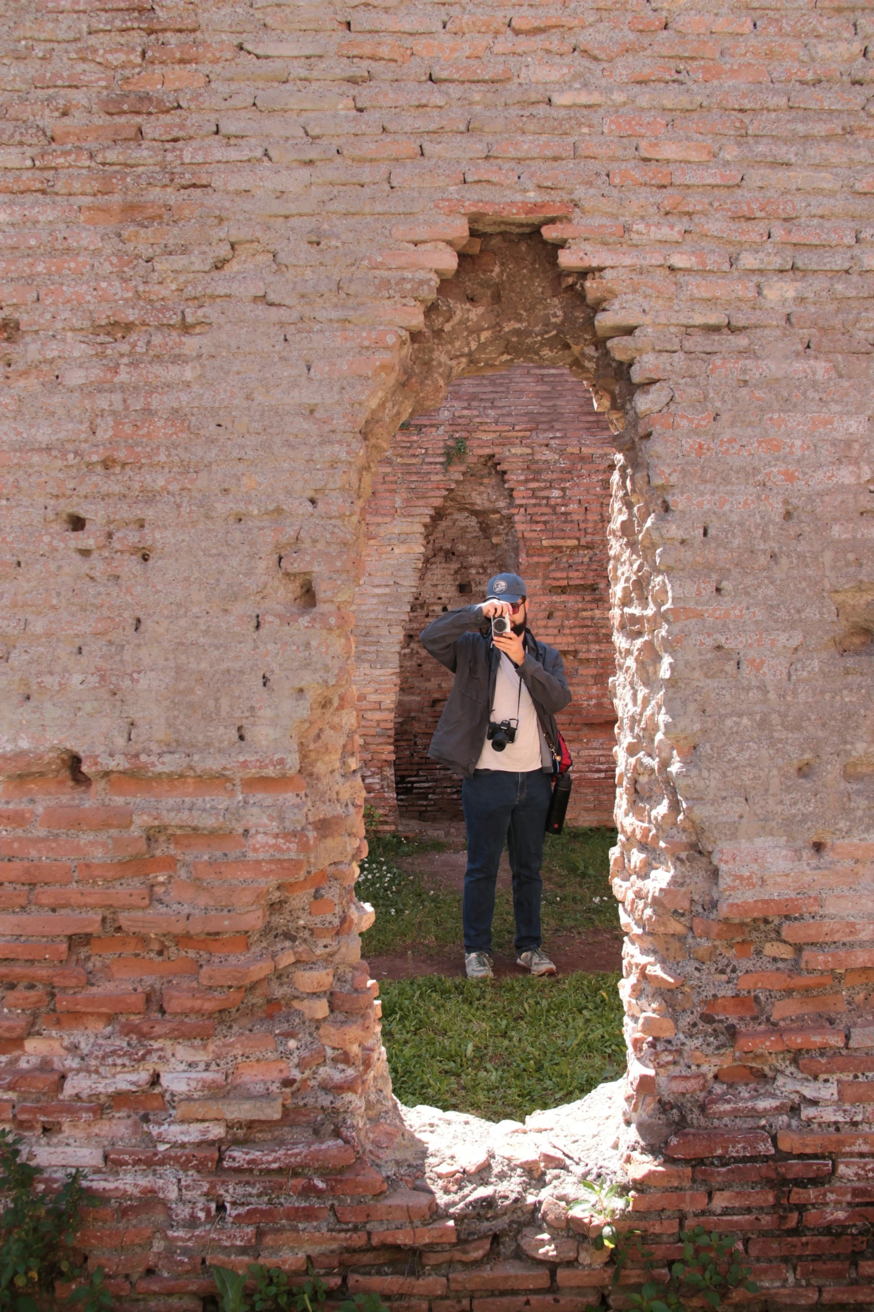 A man standing in a ruined brick doorway, raising a camera to his eye.